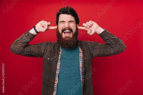 Obraz na płótnie Frustrated bearded man in brown shirt holding fingers in his ears and keeping ey