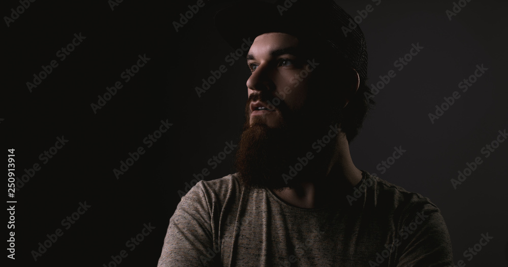 Handsome bearded man in tshirt and hat with crossed arms looking away on darkling background
