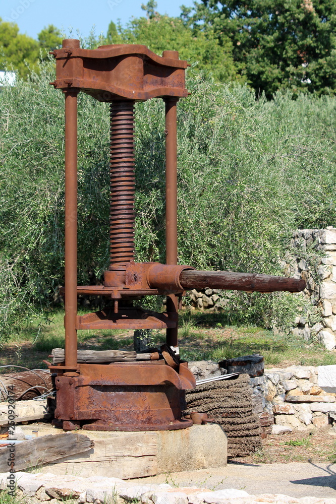 Rusted strong metal vintage retro olive oil press machine mounted on  concrete foundation and used as garden decoration surrounded with  traditional stone wall and olive trees in background on warm sunn Stock