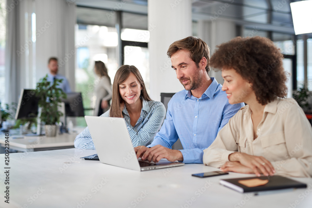 Coworkers sitting at table and solving problem. Man using laptop while two female colleagues looking at laptop and helping him.. Coworkers sitting at table and solving problem. Man using laptop while two female colleagues looking at laptop and helping him..