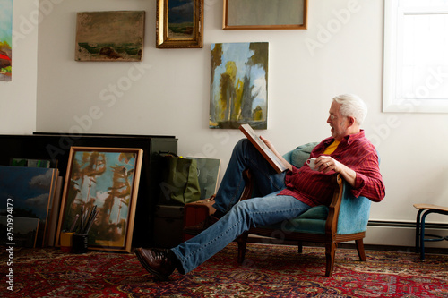 Senior man sitting on armchair in house interior,  looking at artwork,  oil paintings hanging and leaning by wall  