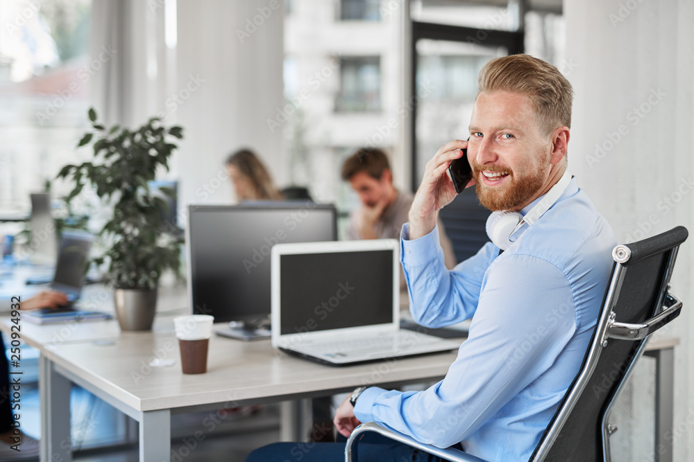 Smiling ginger CEO looking over shoulder and talking on the phone while sitting in modern office. Smiling ginger CEO looking over shoulder and talking on the phone while sitting in modern office.