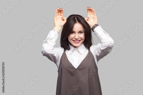 Portrait of funny beautiful young woman in white shirt and brown apron with makeup and brunette hair with bunny ears gesture and looking at camera with toothy smile. studio shot on grey background.