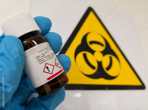 Concept of hazardous material or harmful chemical reagent. Hand with a protective glove holding an amber glass bottle, chemical marked with an attention symbol.