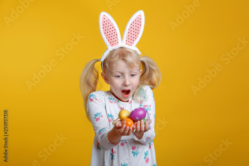 Cute adorable Caucasian blonde girl in white dress with pink  Easter bunny ears holding eggs in studio on yellow background. Funny surprised kid child celebrating traditional Christian holiday