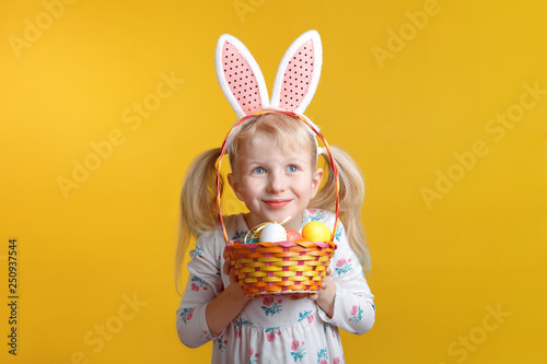 Cute adorable Caucasian blonde girl in white dress wearing pink  Easter bunny ears holding basket with eggs in studio on yellow background. Funny kid child celebrating Christian holiday