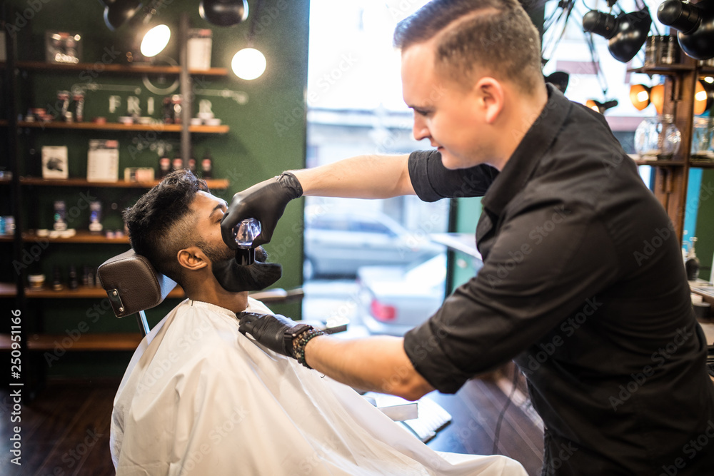 Grooming of real man. Young indian bearded man getting beard haircut at hairdresser while sitting in chair at barbershop Grooming of real man. Young indian bearded man getting beard haircut at hairdresser while sitting in chair at barbershop
