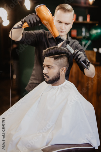 Barber with hairdryer drying and styling hair of client. Barber with hairdryer works on hairstyle for bearded man, barbershop background. Styling concept. Hipster bearded client getting hairstyle.