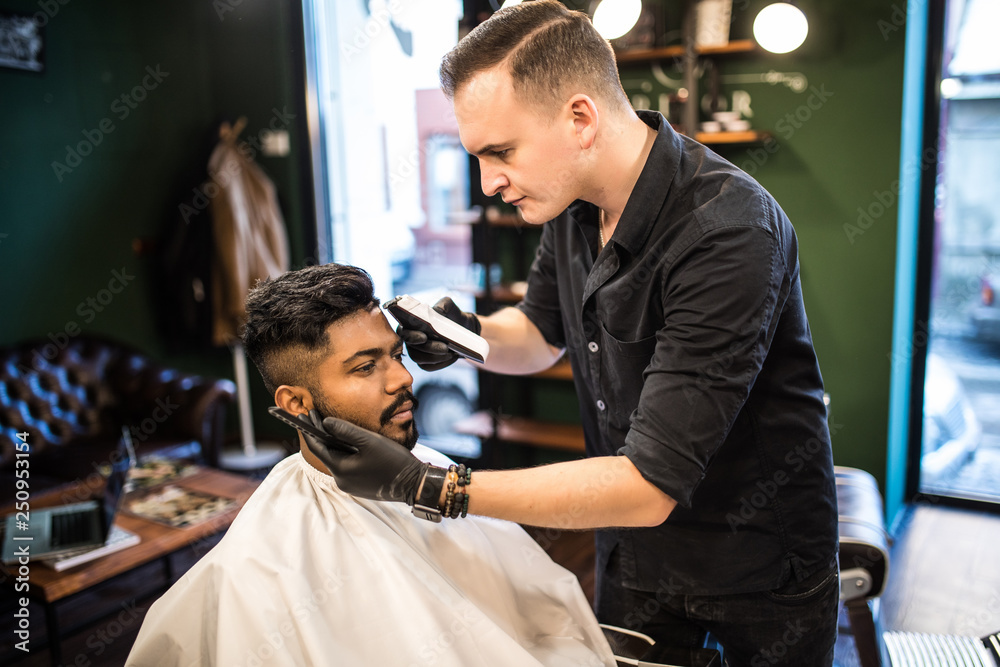 Professional styling. Young bearded man getting haircut by hairdresser with electric razor at barbershop Professional styling. Young bearded man getting haircut by hairdresser with electric razor at barbershop