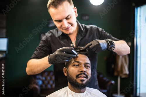 Young man being clipped with professional electric shearer machine in barbershop. Male beauty treatment concept. Indian guy trim beard and mustache at barber shop salon