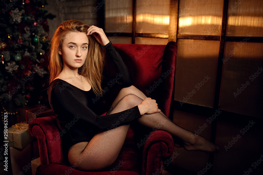 beautiful woman with long brunette bulky hair in dark clothes posing on the red chair in studio with Christmas tree on background beautiful woman with long brunette bulky hair in dark clothes posing on the red chair in studio with Christmas tree on background