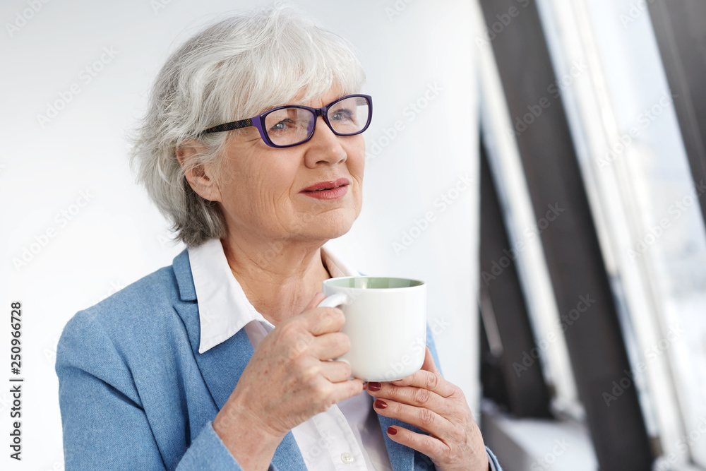 Pensive beautiful female pensioner wearing stylish rectangular eyeglasses and blue jacket holding mug, enjoying aroma of good fresh cappuccino. Gray haired elegant senior woman drinking tea Pensive beautiful female pensioner wearing stylish rectangular eyeglasses and blue jacket holding mug, enjoying aroma of good fresh cappuccino. Gray haired elegant senior woman drinking tea
