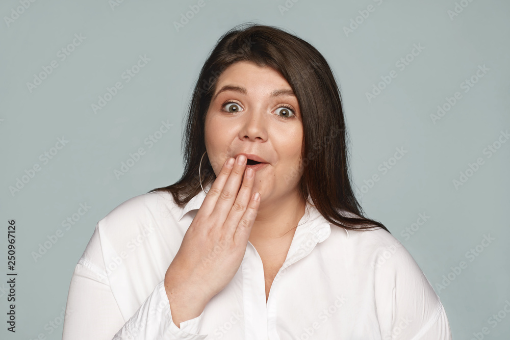 Omg. Human reaction and emotions. Portrait of astonished fascinated young obese Caucasian woman employee with chubby cheeks covering mouth, shocked with unexpected gossip about her colleague Omg. Human reaction and emotions. Portrait of astonished fascinated young obese Caucasian woman employee with chubby cheeks covering mouth, shocked with unexpected gossip about her colleague