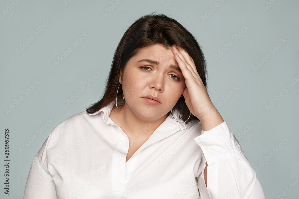 Studio shot of upset frustrated young chubby female employee wearing formal shirt holding hand on her forehead, having mournful look, suffering from headache or hangover after sleepless night Studio shot of upset frustrated young chubby female employee wearing formal shirt holding hand on her forehead, having mournful look, suffering from headache or hangover after sleepless night