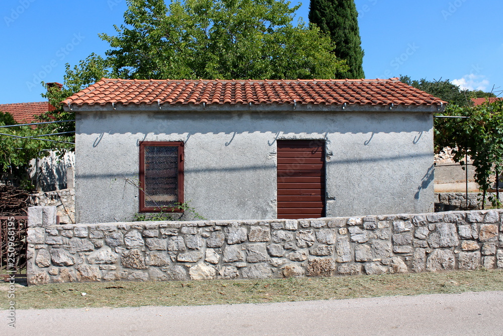 Small backyard unfinished outdoor storage structure with window and wooden door surrounded with traditional stone wall and trees in background on warm sunny day Small backyard unfinished outdoor storage structure with window and wooden door surrounded with traditional stone wall and trees in background on warm sunny day