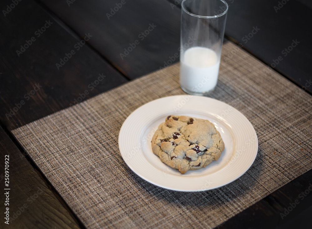 Delicious chocolate chip cookie on a plate and a glass of milk sitting on a table Delicious chocolate chip cookie on a plate and a glass of milk sitting on a table