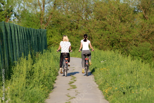 Two beautiful young girls in sport outfit riding their bicycles on paved country path surrounded with high green grass and forest vegetation next to metal fence on warm sunny day
