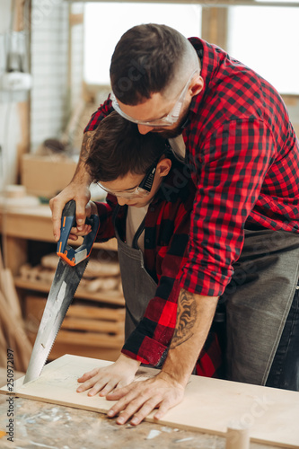 фотографія Father and little son at carpentry workshop standing near table dad hugging boy holding saw together sawing a wooden plank
