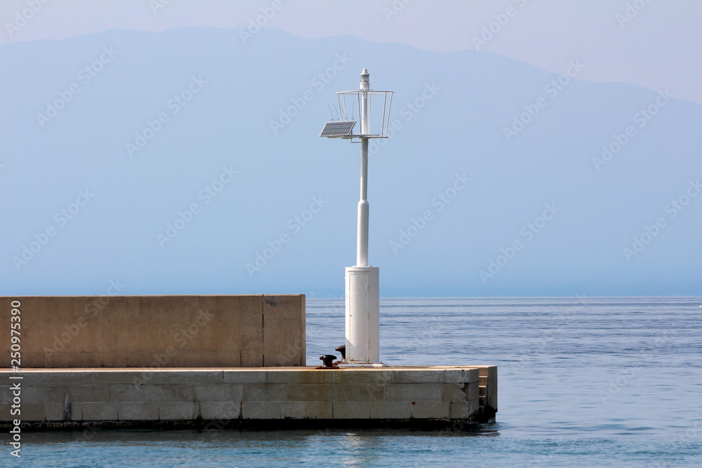 White metal pole with light beacon acting as local lighthouse powered with small solar panel on edge of stone tiles pier surrounded with calm blue sea on warm sunny day