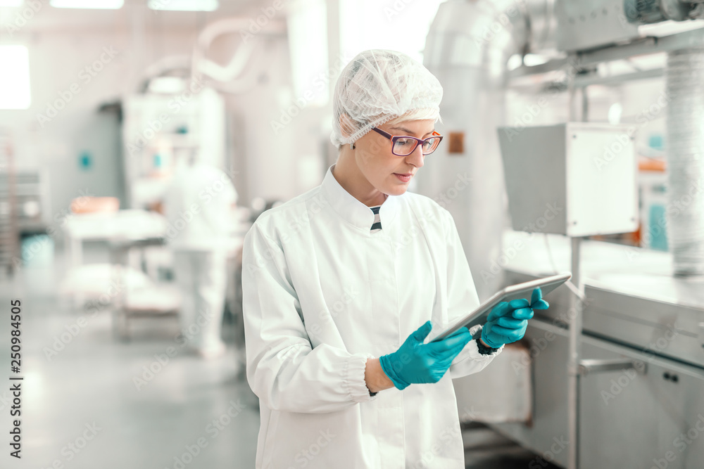 Supervisor in sterile uniform and with eyeglasses using tablet for controlling workflow in food factory. Supervisor in sterile uniform and with eyeglasses using tablet for controlling workflow in food factory.