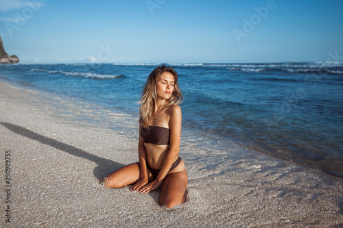 Sexy girl in a brown swimsuit sits on her knees on the beach with white sand, on the background of the ocean.