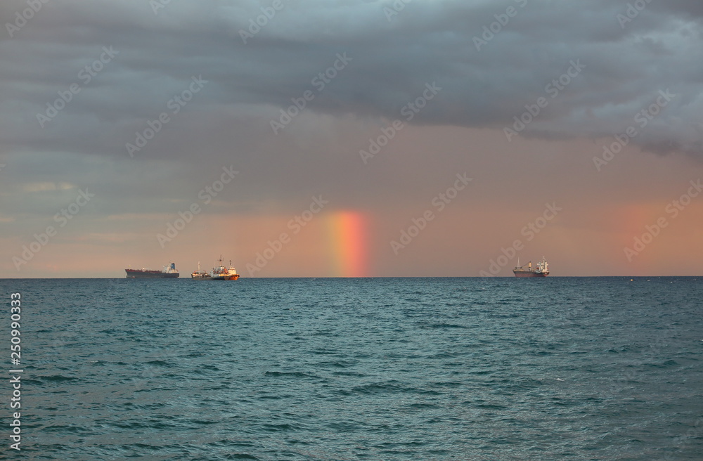 Short rainbow over sea, between three big tankers, under heavy clouds
