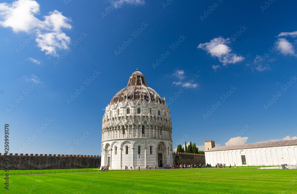 Pisa Baptistery Battistero di Pisa on Piazza del Miracoli Duomo square green grass lawn, city wall, Camposanto cemetery, blue sky with white clouds copy space background in sunny day, Tuscany, Italy
