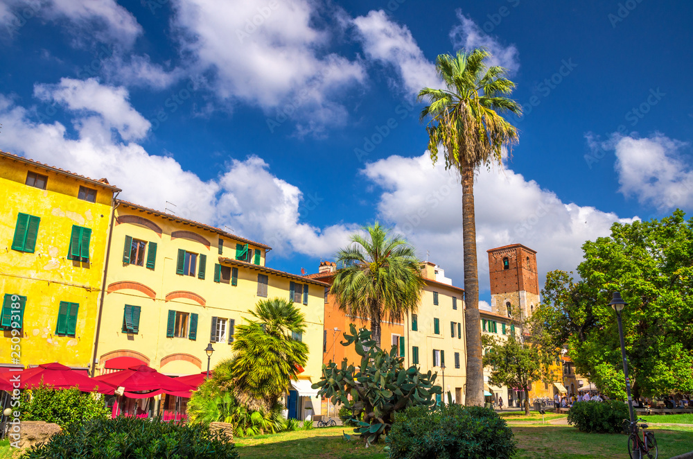 Idee In Circolo park with palms, colorful buildings and bell tower on Piazza Dante Alighieri square in historical centre of Pisa town with blue sky white clouds background in sunny day, Tuscany, Italy
