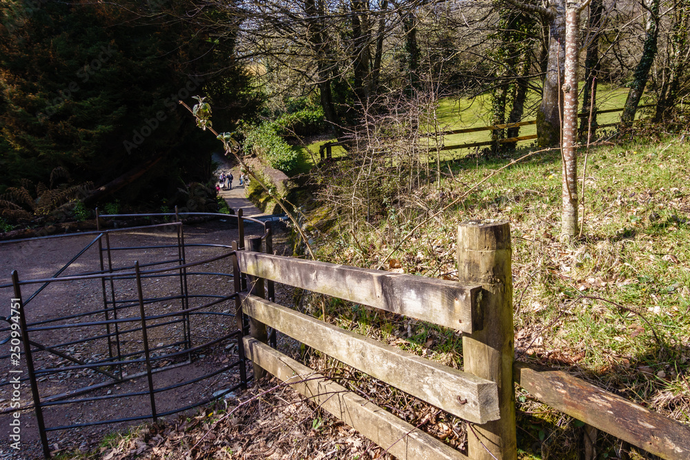 wooden fence and metal fence for entrance to the ancient park; gravelly walk along ornamental greenery and large trees; spring day and trees are leafless