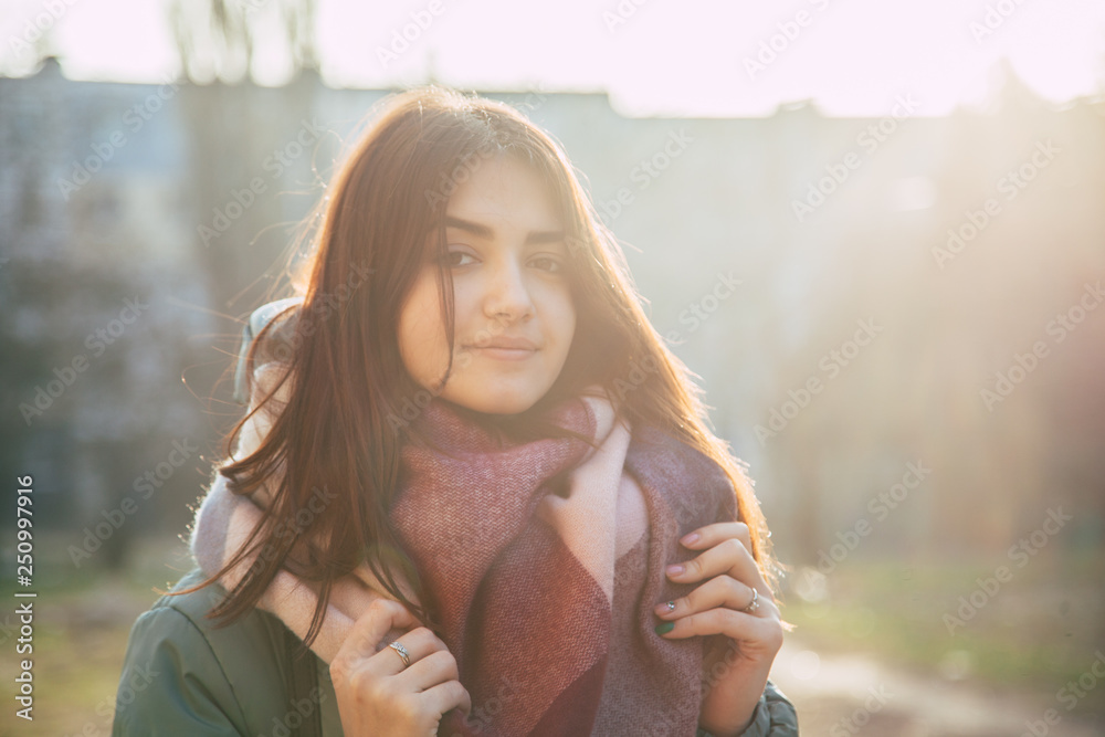 young positive fasion girl standing outdoors in winter coat, touching scarf with two hands in the sunny day
