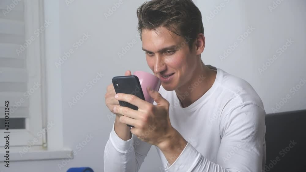 Attractive smiling man using smartphone sending message on social media and drinking coffee indoor