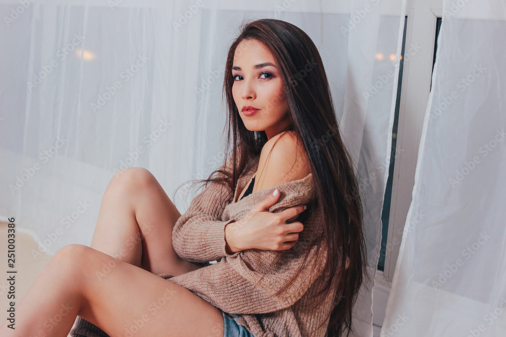 Naklejka premium Candid portrait of sensual smiling asian girl young woman with dark long hair in cozy beige sweater on white curtain background