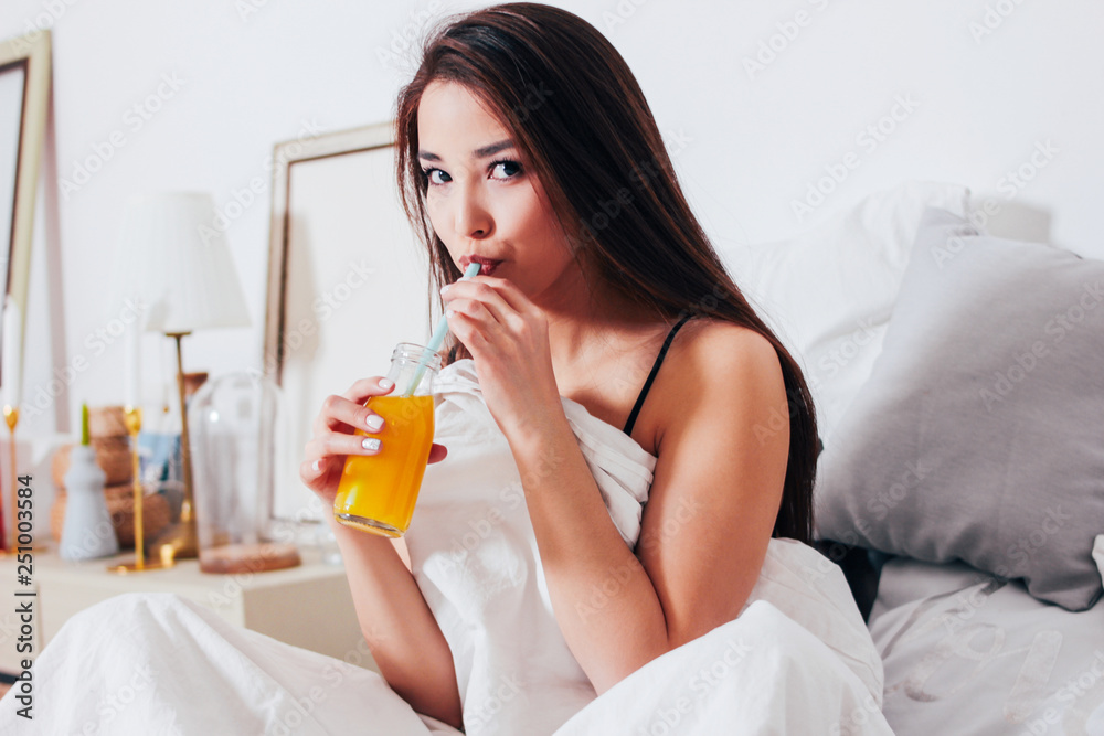 Happy smiling beautiful long hair asian girl young woman sitting in bed with orange juice in jar, healthy cozy Happy smiling beautiful long hair asian girl young woman sitting in bed with orange juice in jar, healthy cozy