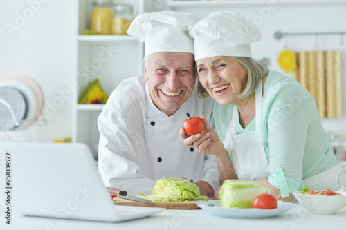 Portrait of senior couple at kitchen cooking with laptop