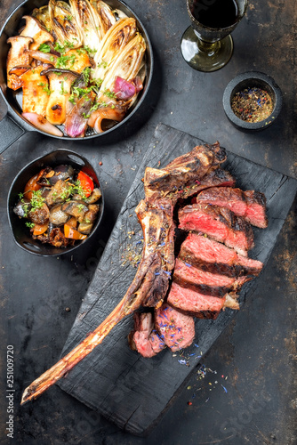 Traditional barbecue dry aged wagyu tomahawk steak sliced with ratatouille and fried mushroom chicory as top view on a rustic carbonized wooden board