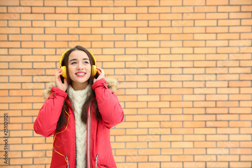 Beautiful young woman listening to music with headphones against brick wall. Space for text