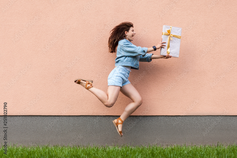 Φωτογραφία Full length side view of happy excited beautiful woman in casual jeans denim style in summertime standing near sandybrown wall, holding gift box and jumping