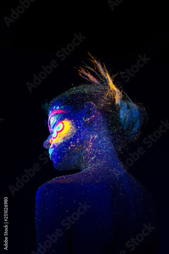 Blue alien girl stands in the dark, view from the back. Ultraviolet makeup and light.