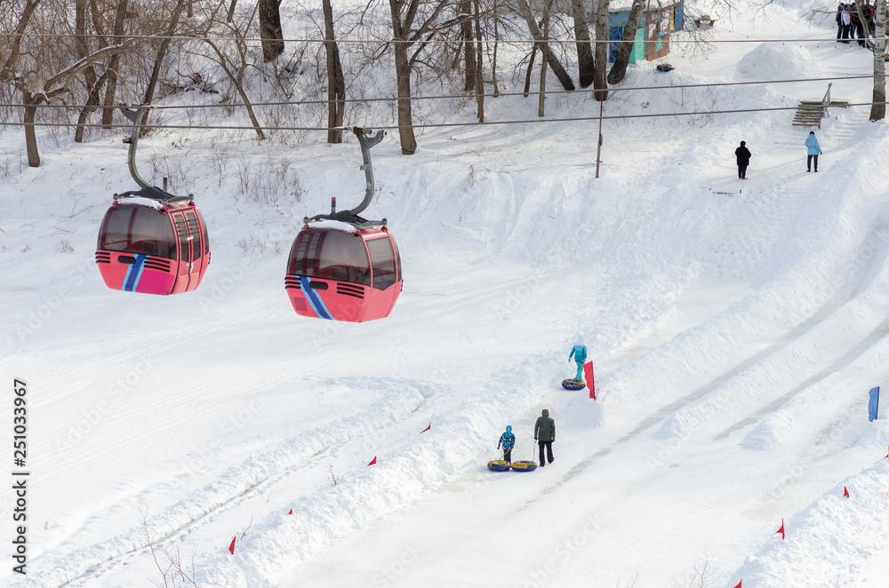 The cabins of funiculars over the frozen river and ice slides. Orenburg, Russia - February, 21, 2019: Ural River in winter. Cable car and skiing from an ice slide in the Zauralny grove