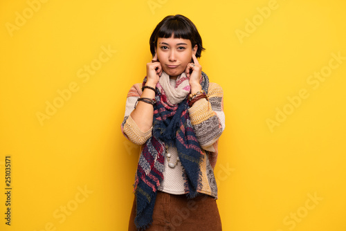 Young hippie woman over yellow wall covering ears with hands. Frustrated expression