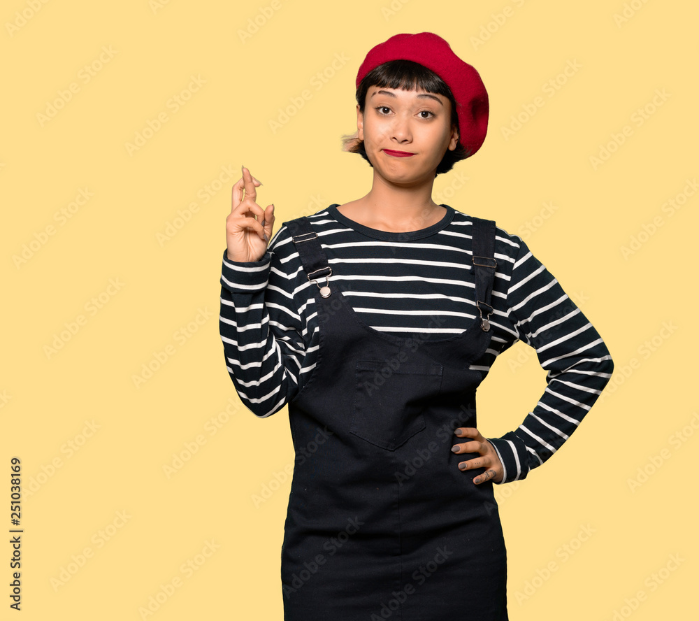 Young woman with beret with fingers crossing and wishing the best over yellow background Young woman with beret with fingers crossing and wishing the best over yellow background