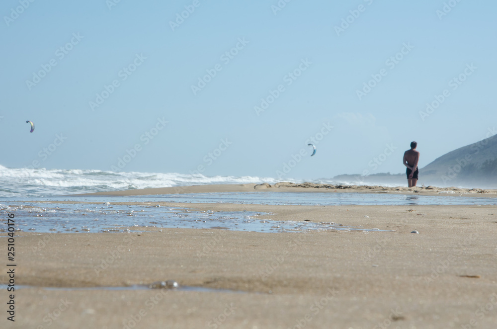 Close-up. Sea beach with the oncoming wave. Wet sand On the average plan is a man, he looks into the distance. In the background are the kitesurf sails. Sunny windy day.