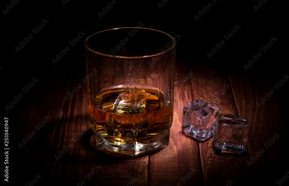 A glass of whisky and some ice cubes on the wooden table A glass of whisky and some ice cubes on the wooden table