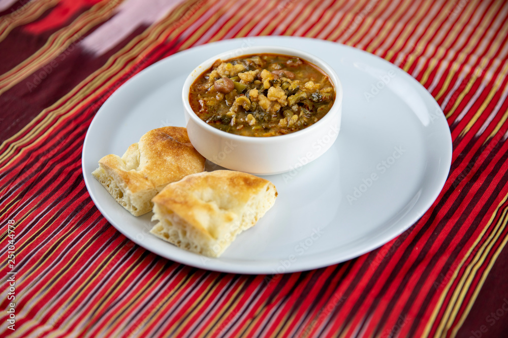 A bowl of hearty soup with chunks of vegetables and grains, served with pieces of crusty bread on a white plate, accompanied by a wooden utensil, set against a vibrant red and gold striped background. A bowl of hearty soup with chunks of vegetables and grains, served with pieces of crusty bread on a white plate, accompanied by a wooden utensil, set against a vibrant red and gold striped background.