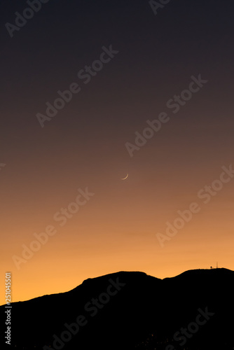 Beautiful sunset over the mountain silhouette with the moon on the clear sky, street lights of inhabited places visible on the mountain slopes, vertical landscape