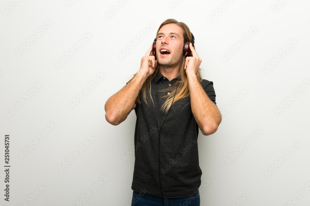 Blond man with long hair over white wall listening to music with headphones Blond man with long hair over white wall listening to music with headphones