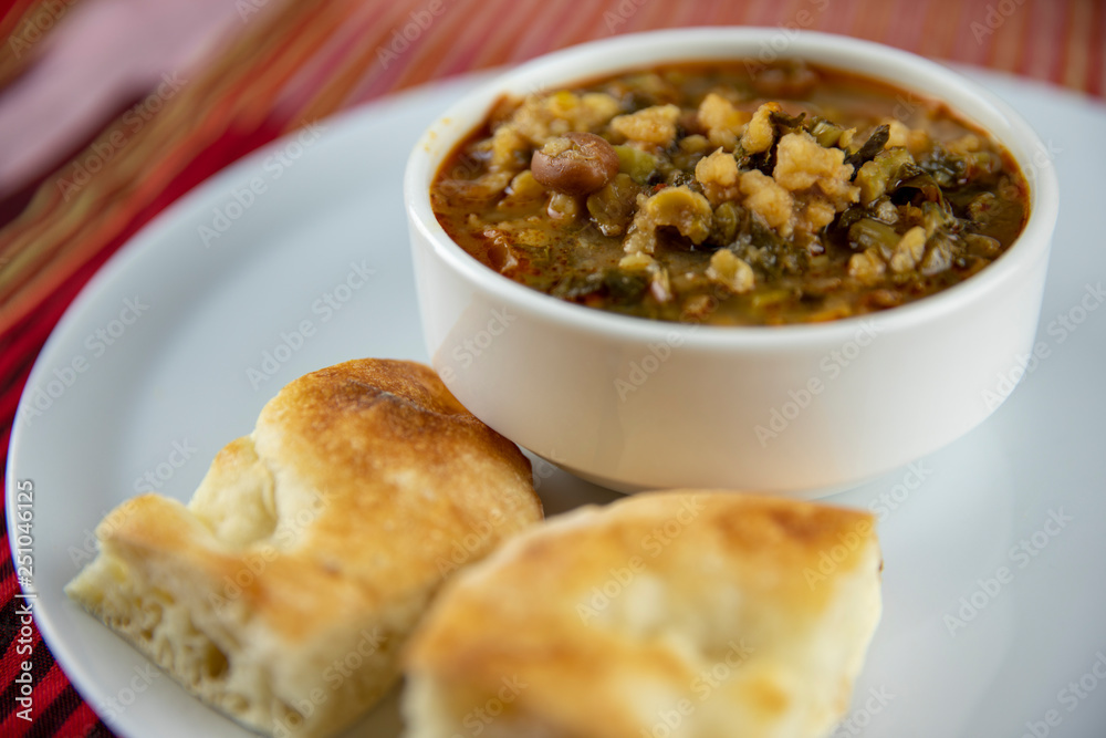 A bowl of hearty soup with chunks of vegetables and grains, served with pieces of crusty bread on a white plate, accompanied by a wooden utensil, set against a vibrant red and gold striped background. A bowl of hearty soup with chunks of vegetables and grains, served with pieces of crusty bread on a white plate, accompanied by a wooden utensil, set against a vibrant red and gold striped background.