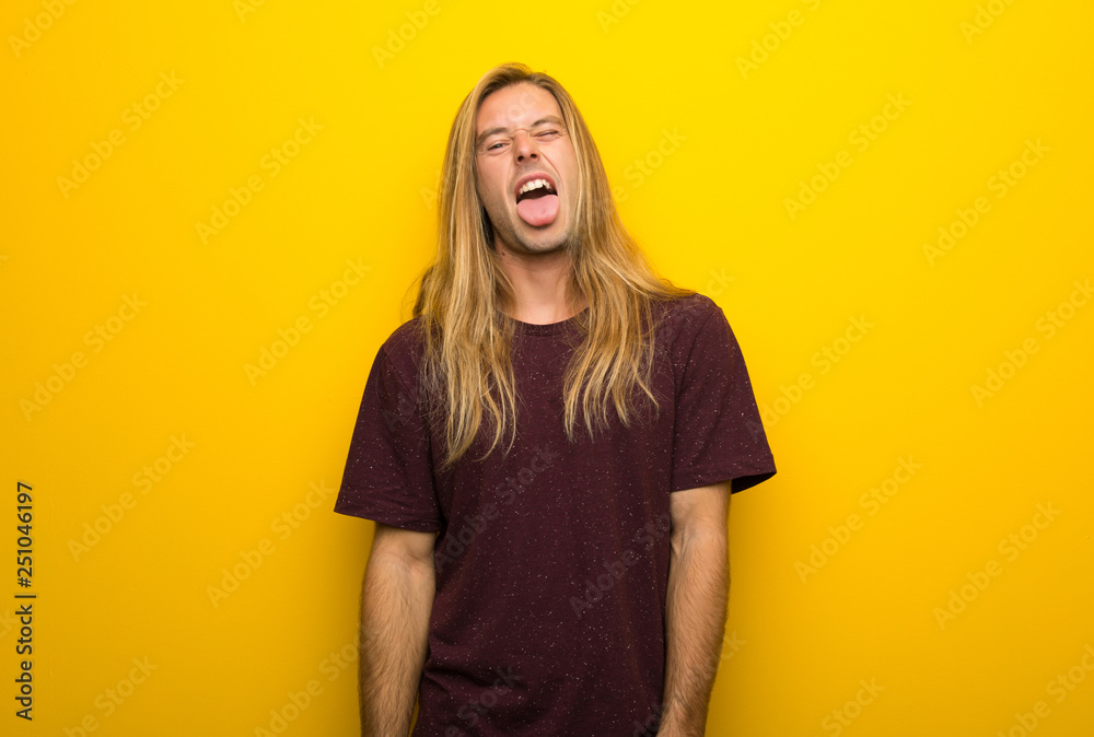Blond man with long hair over yellow wall showing tongue at the camera having funny look Blond man with long hair over yellow wall showing tongue at the camera having funny look
