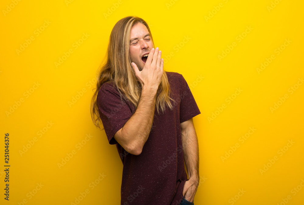 Blond man with long hair over yellow wall yawning and covering wide open mouth with hand Blond man with long hair over yellow wall yawning and covering wide open mouth with hand