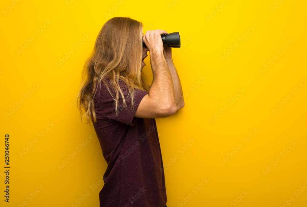 Blond man with long hair over yellow wall and looking in the distance with binoculars Blond man with long hair over yellow wall and looking in the distance with binoculars